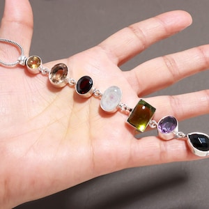 Può includere: Una collana a catena in argento con una serie di sette pietre preziose multicolori di varie forme, inclusi tagli rotondi, ovali, rettangolari e a goccia. Le pietre presentano tonalità di giallo, marrone, rosso scuro, bianco, verde, viola e nero.