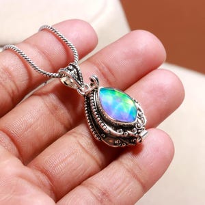May include: A silver pendant necklace featuring an oval opal gemstone with iridescent green, blue, and purple hues. The pendant has an ornate border and hangs from a fine silver chain. The necklace is a piece of jewellery.