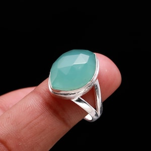 Pode incluir: Um anel de prata com uma grande pedra preciosa azul clara facetada em forma de gota. O anel tem uma banda simples de haste dividida. A pedra preciosa é definida em uma configuração de moldura. Mostrado em um fundo preto.