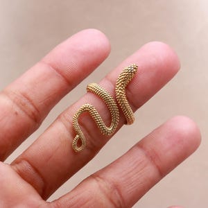 Puede incluir: Un anillo de serpiente dorado con un patrón texturizado en forma de escamas. El anillo se enrolla alrededor de un dedo, con la cabeza y la cola de la serpiente formando los extremos. Es una joya.