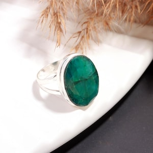 May include: A silver ring featuring a large, oval-cut emerald gemstone. The emerald is a vibrant green color and is set in a polished silver band. The ring is displayed on a white surface.