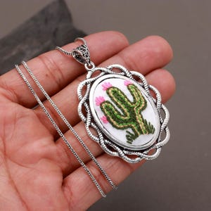 Op de afbeelding: Een ovale hangerketting met een geborduurde groene cactus met roze bloemen op een witte stoffen achtergrond, gevat in een decoratieve zilverkleurige metalen omlijsting met een gedraaide rand en een bijpassende zilverkleurige ketting.