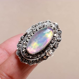 Könnte beinhalten: Silberring mit einem ovalen Opal-Edelstein. Der Opal zeigt ein Farbspiel, darunter Rosa, Blau und Gelb. Der Ring hat ein aufwendiges Filigran-Design mit kleinen, kreisförmigen Verzierungen um den Opal.