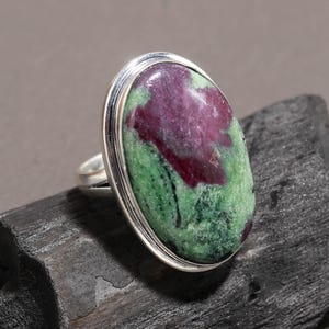 May include: A silver ring featuring a large, oval-shaped cabochon stone with a mottled appearance. The stone displays a vibrant mix of deep magenta-red patches set against a textured, moss-green background, held securely in a smooth metal bezel.