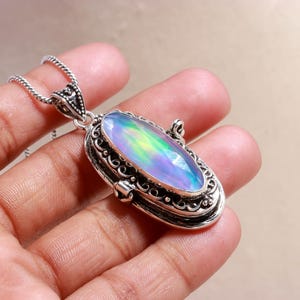 Könnte beinhalten: Silberfarbene Halskette mit einem ovalen Anhänger mit einem schillernden Opal-Edelstein. Der Anhänger ist mit aufwendigen Schnörkeln und einem dekorativen Bügel versehen. Der Opal zeigt ein Farbspektrum, darunter Blau, Grün und Gelb.