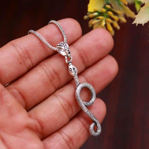 Può includere: Un ciondolo a forma di serpente color argento appeso a una catenina abbinata. Il ciondolo presenta un corpo di serpente dettagliato e testurizzato che si avvolge in un piccolo anello alla base. La contromaglia superiore ha un motivo inciso.