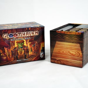 Puede incluir: Dos cajas para el juego de mesa "Gloomhaven: Buttons & Bugs". La caja del juego presenta una colorida ilustración de fantasía con el título del juego. La otra caja tiene un diseño de vetas de madera y contiene componentes del juego.