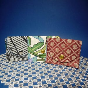 Op de afbeelding: Drie kleine stoffen zakjes met drukknoopsluitingen. Eén is zwart-wit gestreept, een ander heeft een bloemenpatroon met groene bladeren en de derde is rood met een ruit- en bloemenpatroon. De zakjes worden op een blauw oppervlak gepresenteerd.