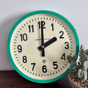 Può includere: Un orologio da parete rotondo con un quadrante color crema e numeri neri. L'orologio ha un bordo verde e lancette nere. L'orologio è accanto a una piccola pianta in vaso.