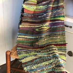 May include: A vibrant, hand-woven textile with a textured, layered design. It displays horizontal stripes in green, brown, red, and blue. The edges are fringed. The textile is draped over a wooden chair with a woven seat. The piece is reminiscent of a throw.