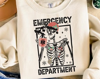 Op de afbeelding: Crèmekleurige sweatshirt met de tekst "EMERGENCY DEPARTMENT" in zwart. De afbeelding toont een skelet met een kerstmuts en een koffiekopje met een medisch symbool. De ribbenkast van het skelet is versierd met kerstversieringen.