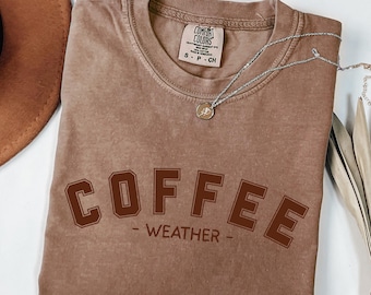 Könnte beinhalten: Ein braunes Comfort Colors T-Shirt mit den Worten "COFFEE WEATHER" in dunklerem Braun aufgedruckt. Eine silberne Halskette mit einem kleinen Anhänger liegt über dem Shirt. Ein brauner Hut ist in der oberen linken Ecke zu sehen.