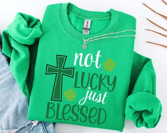 May include: A green sweatshirt with the words "not LUCKY just BLESSED" in white and green lettering. The design includes a cross and shamrocks. A silver necklace is draped across the top of the sweatshirt.