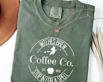 Puede incluir: Camiseta verde oliva con el texto "Witches Brew Coffee Co." en un diseño circular. El diseño incluye una bruja en una escoba y el texto "Stop in for a spell". También se ven un collar dorado y un sombrero marrón.