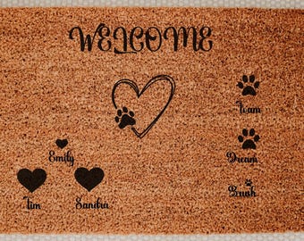 May include: A rectangular doormat with a brown coir surface. The word "welcome" is arched across the top in black script. A heart with a paw print is in the center. Names and paw prints are below.
