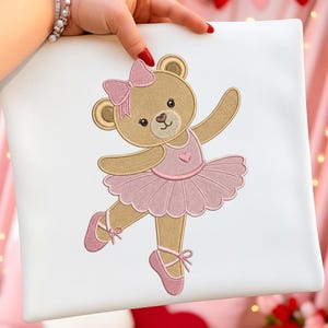 Könnte beinhalten: Ein weißes quadratisches Kissen mit einem gestickten Design eines Ballerina-Teddybären. Der Bär ist hellbraun mit einer rosa Schleife, einem Tutu und Ballettschuhen. Das Design enthält ein Herz auf der Brust des Bären.