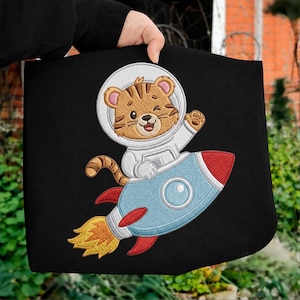 May include: Black tote bag featuring an embroidered cartoon tiger in a spacesuit riding a blue and red rocket. The tiger is winking and waving. The rocket has a yellow flame. The design is on a black background.