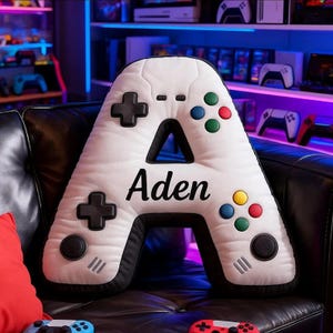 May include: A white letter A-shaped pillow featuring black and colored gaming controller button graphics. The center of the pillow displays the name Aden in black script. The design includes a black directional pad, two black joysticks, and red, blue, yellow, and green action buttons.