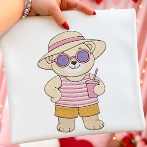 May include: Embroidered white tote bag featuring a cartoon bear wearing sunglasses, a sun hat, and a striped shirt. The bear is holding a pink drink with a straw and wearing yellow shorts. The design is in shades of tan, pink, and yellow.