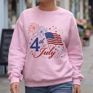 May include: A light pink crewneck sweatshirt featuring a graphic design with the text 4th of July in blue and red. The design includes a waving American flag, red and blue firework bursts, and small star accents scattered across the chest area.