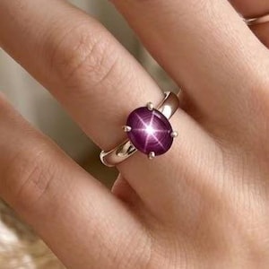 Könnte beinhalten: Ein silberner Ring mit einem großen, ovalen, tiefrosa Edelstein. Der Edelstein weist ein sternförmiges Muster auf. Ein schlichter silberner Bandring ist ebenfalls zu sehen. Der Schmuck wird auf einer Hand präsentiert.