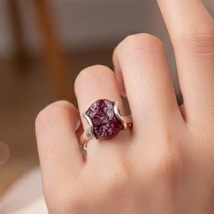 Könnte beinhalten: Ein Silberring mit einem großen, roh geschnittenen, tiefroten Edelstein. Der Stein ist unregelmäßig geformt und in einem minimalistischen Silberband gefasst. Der Ring wird an einem Finger gezeigt, wodurch das Design und die Farbe des Schmucks hervorgehoben werden.
