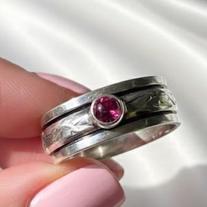 May include: A silver ring with a rotating band and a round, magenta-colored gemstone. The ring has an engraved floral pattern on the outer band. The gemstone is set in a bezel setting. The ring is held by a person with pink nail polish.