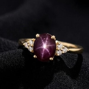 Könnte beinhalten: Ein goldener Ring mit einem großen, ovalen Sternrubin-Edelstein. Der Rubin hat eine tiefe burgunderrote Farbe mit einem sechsstrahligen Sternmuster. Kleine, klare Edelsteine sind auf beiden Seiten des Rubins gesetzt. Der Ring ist vor einem schwarzen Hintergrund platziert.