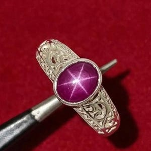Könnte beinhalten: Ein silberner Ring mit einem ovalen, tiefrosa Edelstein, der ein sechszackiges Sternmuster aufweist. Das Band des Rings weist aufwendige, gravierte Details auf. Der Ring wird von einem Werkzeug vor rotem Hintergrund gehalten.