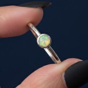 Könnte beinhalten: Ein zarter Silberring mit einem runden, schillernden Opal-Edelstein. Der Opal zeigt eine Mischung aus grünen, gelben und weißen Farbtönen. Der Ring hat ein dünnes, poliertes Band und ein minimalistisches Design.
