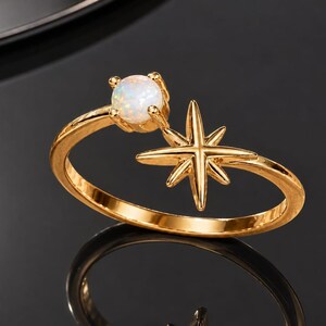 Könnte beinhalten: Ein goldfarbener offener Ring mit einem runden, schillernden weißen Opalstein an einem Ende und einem mehrzackigen Sternmotiv am anderen. Das Band ist poliert und reflektierend und zeigt ein himmlisch inspiriertes Design mit glatter Oberfläche.