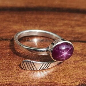 May include: A silver ring featuring a large, oval, deep red gemstone. The gemstone displays a star-like pattern. The ring band has a simple, open design with etched lines on the end.