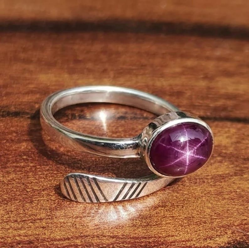 May include: A silver ring featuring a large, oval, deep red gemstone. The gemstone displays a star-like pattern. The ring band has a simple, open design with etched lines on the end.