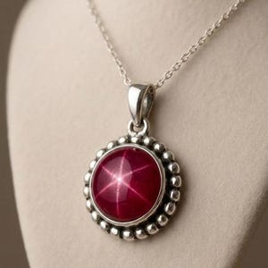 Könnte beinhalten: Eine silberne Anhänger Halskette mit einem runden, tiefroten Edelstein mit sternförmigem Muster. Der Edelstein ist von einem silbernen Perlenrand umgeben und hängt an einer feinen Silberkette. Der Schmuck wird vor einem neutralen Hintergrund präsentiert.