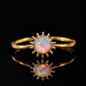 Könnte beinhalten: Ein goldener Ring mit einem zentralen, runden Opal-Cabochon, der ein lebhaftes, irisierendes Farbspiel aufweist. Der Opal ist von einer sonnenförmigen Fassung mit dünnen, strahlenförmigen Metallspitzen umgeben, die vom Rahmen ausgehen.