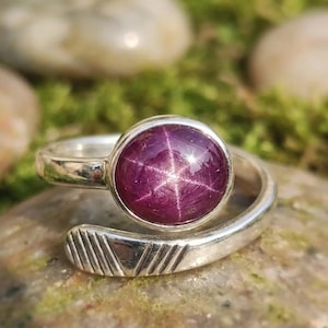 Könnte beinhalten: Ein silberner Ring mit einem großen, runden, tiefroten Edelstein mit sternförmigem Muster. Der Ring hat ein offenes Design mit einem geometrischen Muster am Band. Der Ring ist vor einem natürlichen, moosigen Hintergrund platziert.