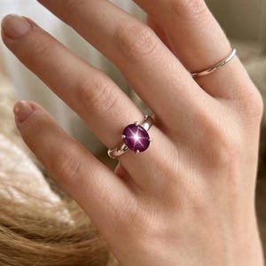 Puede incluir: Un anillo de plata con una gema ovalada de color rojo intenso. La gema tiene un efecto de estrella en el centro. Tambi&eacute;n se ve un segundo anillo de plata m&aacute;s fino. Las joyas se muestran en una mano.