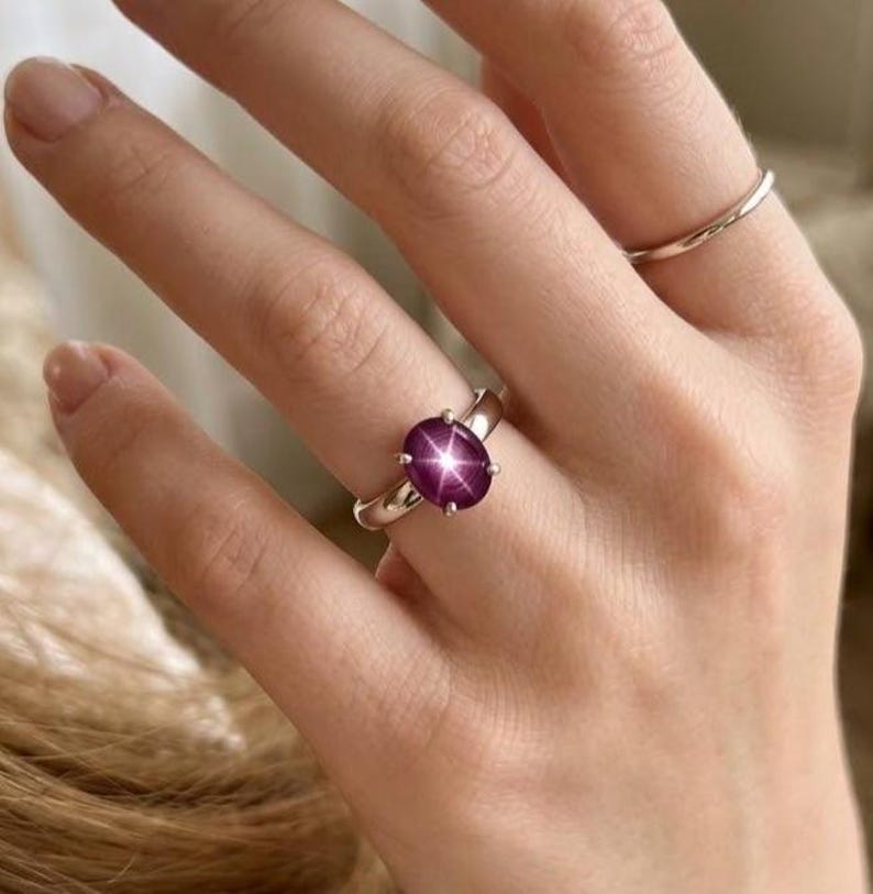 Puede incluir: Un anillo de plata con una gema ovalada de color rojo intenso. La gema tiene un efecto de estrella en el centro. Tambi&eacute;n se ve un segundo anillo de plata m&aacute;s fino. Las joyas se muestran en una mano.