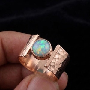Könnte beinhalten: Ein breiter Ring mit einem runden Opal-Cabochon in der Mitte, der irisierende grüne und orangefarbene Blitze aufweist. Der Ring besteht aus einem metallischen Material in Roségold-Optik mit einer texturierten, gehämmerten Oberfläche am Ringband.