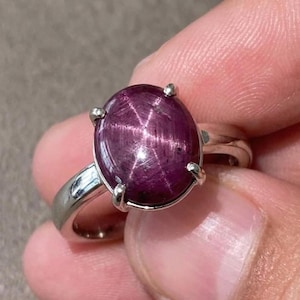 Könnte beinhalten: Ein silberner Ring mit einem großen, ovalen, tiefroten Edelstein mit sternförmigem Muster. Der Edelstein wird von vier silbernen Krappen gehalten. Der Ring hat ein schlichtes, poliertes Silberband.