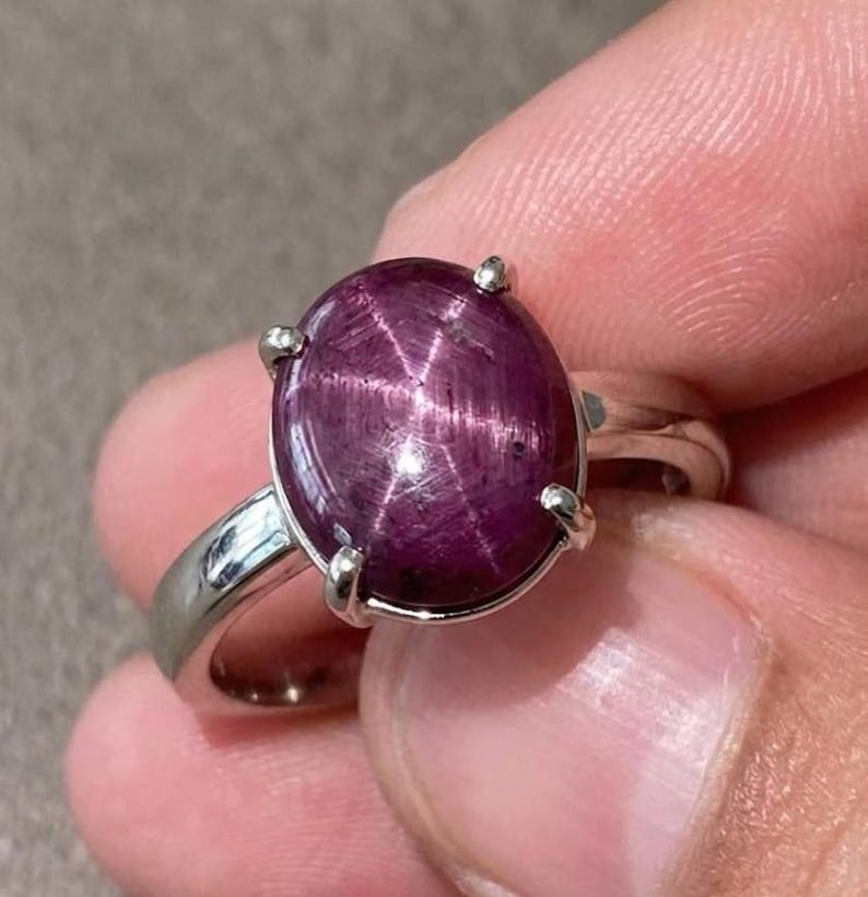 May include: A silver ring with a large, oval, deep red gemstone displaying a star-like effect. The gemstone is secured by four silver prongs. The ring has a simple, polished silver band.