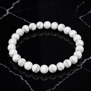 Könnte beinhalten: Ein kreisförmiges Stretch-Armband bestehend aus glatten, runden weißen Perlen mit grauer Maserung. Die Perlen sind einheitlich groß und in einer durchgehenden Schleife angeordnet, was ein schlichtes und klassisches Design für das Handgelenk ergibt.