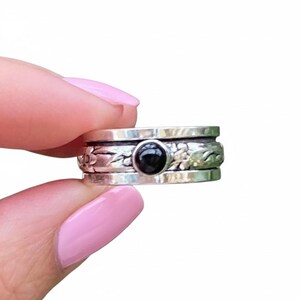Könnte beinhalten: Ein silberfarbener Spinner-Ring mit einem schwarzen Edelstein in der Mitte. Der Ring hat ein Blumenmuster auf dem äußeren Band. Der Ring wird zwischen zwei Fingern mit rosa Nagellack gehalten.