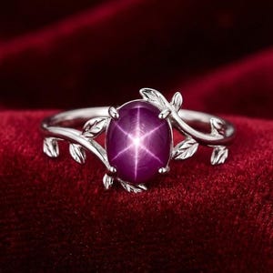 Könnte beinhalten: Ein silberner Ring mit einem großen, ovalen Sternrubin-Edelstein. Das Ringband ist mit filigranen Blatt- und Rankendetails verziert, die ein elegantes Design schaffen. Der Rubin zeigt einen Sechs-Strahl-Stern-Effekt.