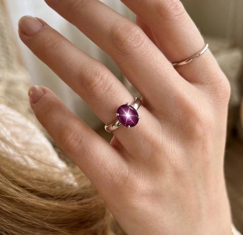 Puede incluir: Un anillo de plata con una gema rosa oscuro ovalada y grande. La gema tiene un patr&oacute;n en forma de estrella. Tambi&eacute;n se ve un anillo de plata liso. La joya se muestra en una mano. El color de la gema es rosa intenso.