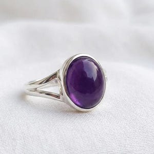 Könnte beinhalten: Ein Silberring mit einem großen, ovalen, tiefvioletten Amethyst-Edelstein. Der Ring hat ein geteiltes Schaftdesign mit zwei parallelen Bändern, die sich an der Basis des Edelsteins treffen. Der Amethyst ist in einer polierten Silberfassung gefasst.