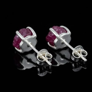 Könnte beinhalten: Ein Paar Ohrstecker mit rohen, tiefroten Rubin-Edelsteinen, die in silberfarbenem Metall gefasst sind. Die Ohrringe haben einen Stecker und einen Butterfly-Verschluss. Der Hintergrund ist schwarz und hebt den Schmuck hervor.