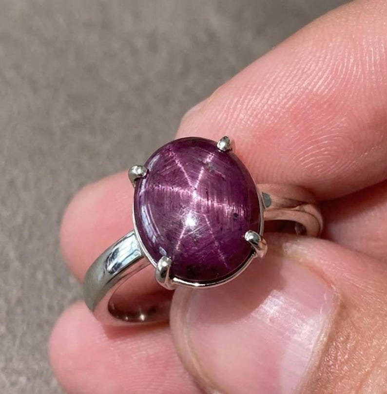 May include: A silver ring with a large, oval, deep purple gemstone displaying a star pattern. The ring has a polished silver band, and the gemstone is secured by four silver prongs.