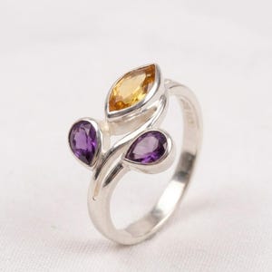 Könnte beinhalten: Ein Silberring mit drei Edelsteinen. Der Ring hat ein blattähnliches Design mit einem gelben Stein im Marquiseschliff und zwei birnenförmigen lila Steinen. Der Ring ist vor einem weißen Hintergrund platziert.