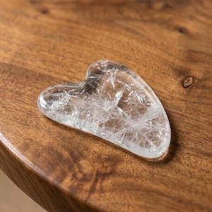 May include: A clear, heart-shaped gua sha tool made of crystal. The tool is resting on a wooden surface. The crystal has internal fractures, giving it a textured appearance. The gua sha tool is used for facial massage and skincare.